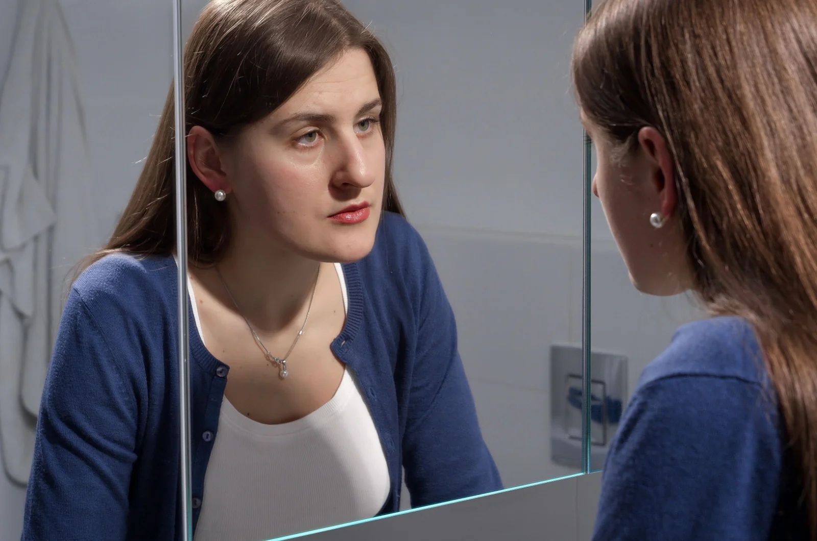 Verärgerte junge Frau steht im Badezimmer und betrachtet ihr Spiegelbild im Spiegel