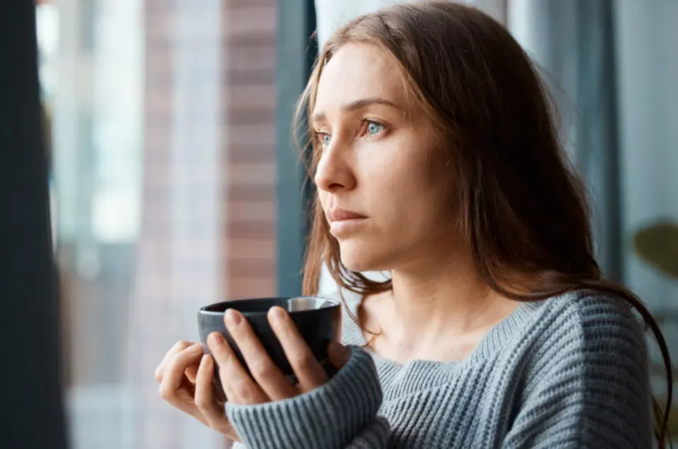 unglueckliche junge Frau trinkt Tee, waehrend sie aus dem Fenster schaut