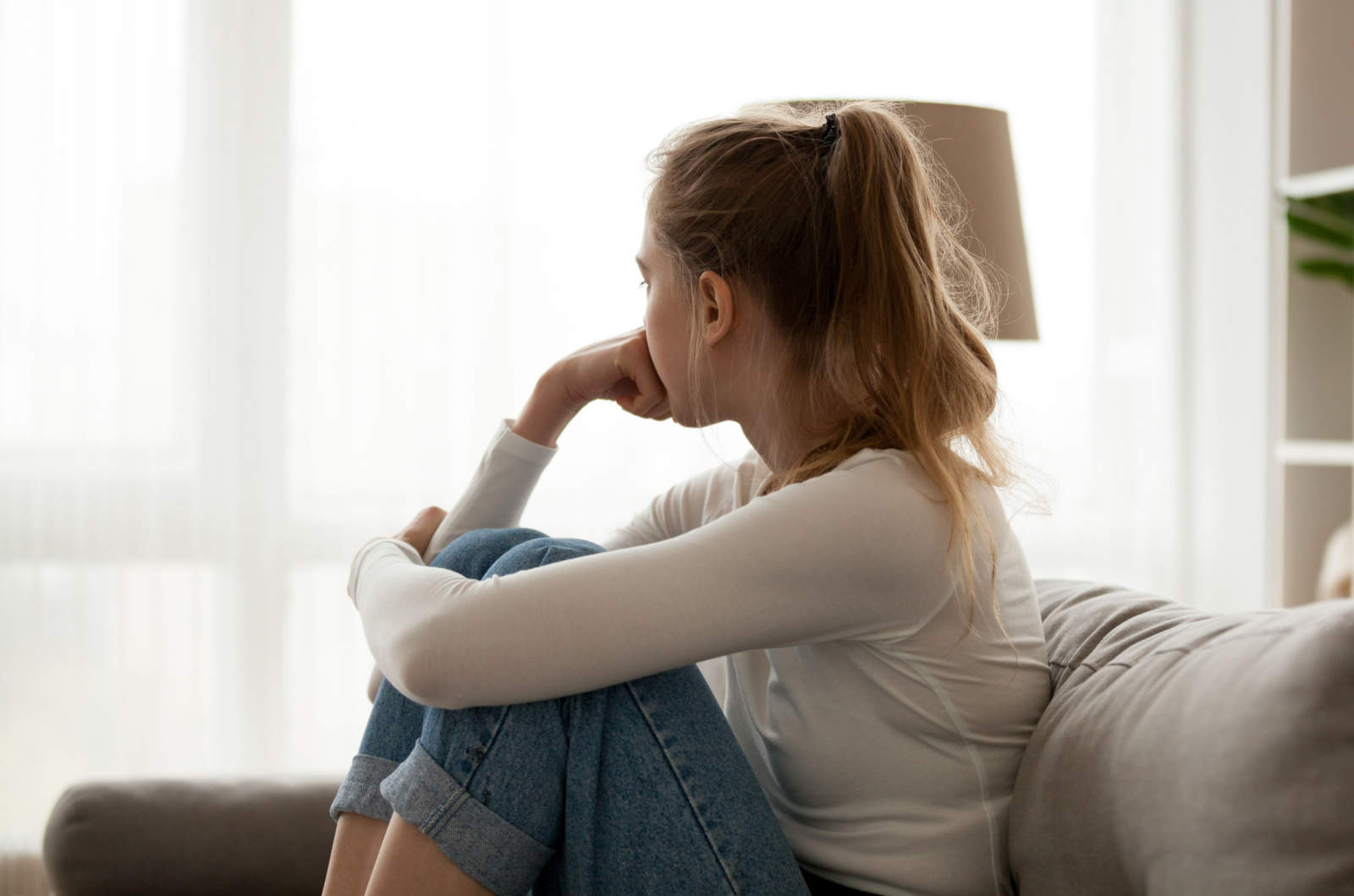 Seitenansicht einer jungen Frau, die zu Hause auf der Couch sitzt und zum Fenster schaut