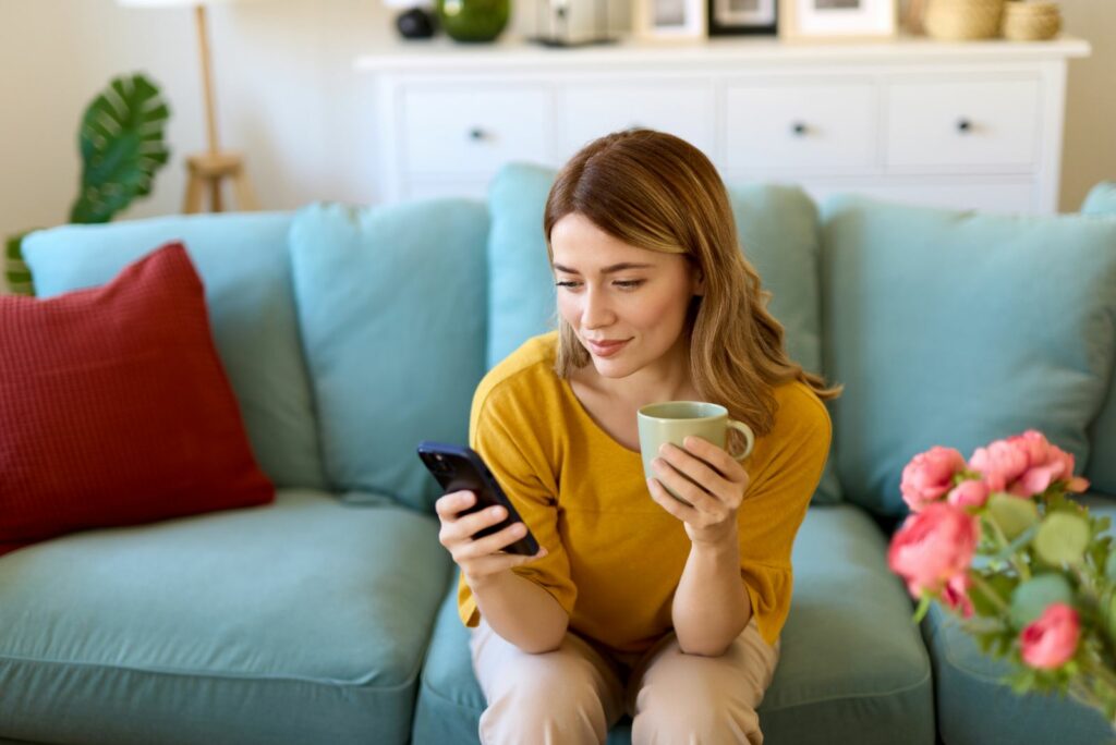 Das Maedchen sitzt mit einer Tasse Kaffee auf der Couch und tippt auf einem Mobiltelefon