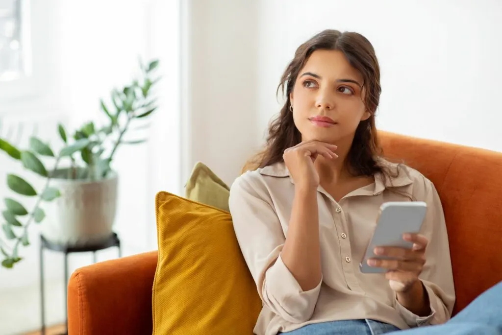 Ein schoenes Maedchen sitzt auf einem Sofa und haelt ein Mobiltelefon in der Hand