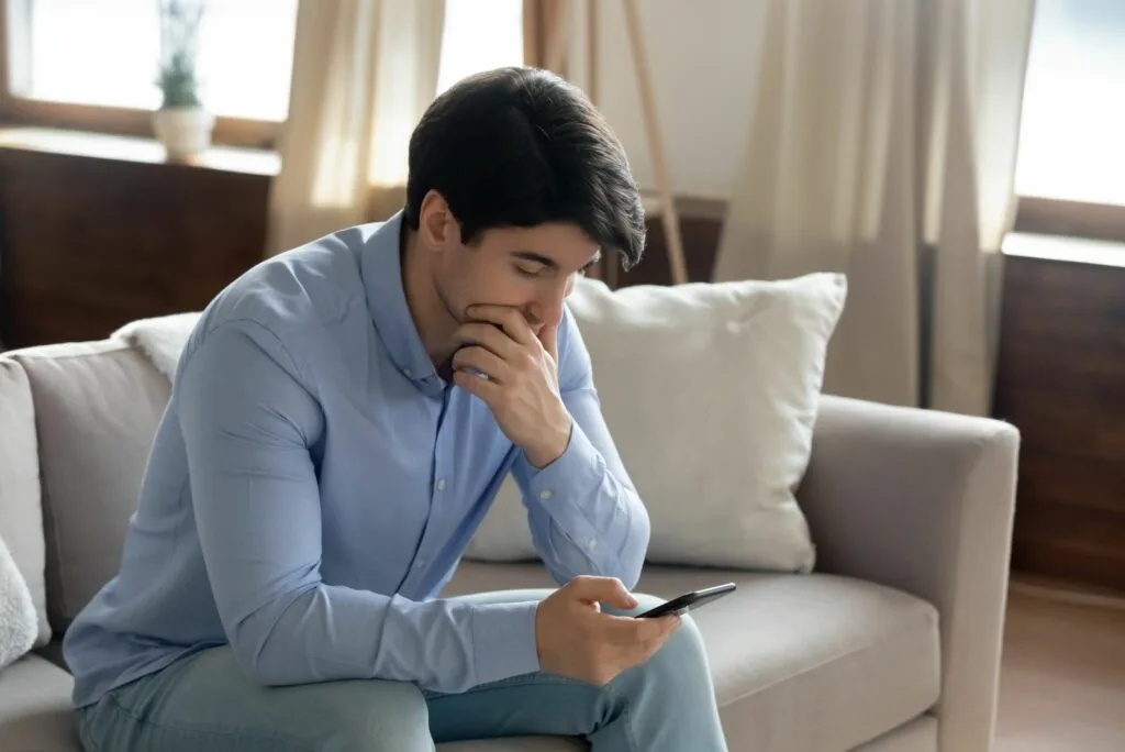 Nachdenklicher Mann sitzt auf dem Sofa und haelt ein Mobiltelefon in der Hand